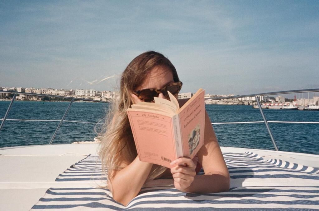 A person lying on a striped towel on a boat, reading a book.