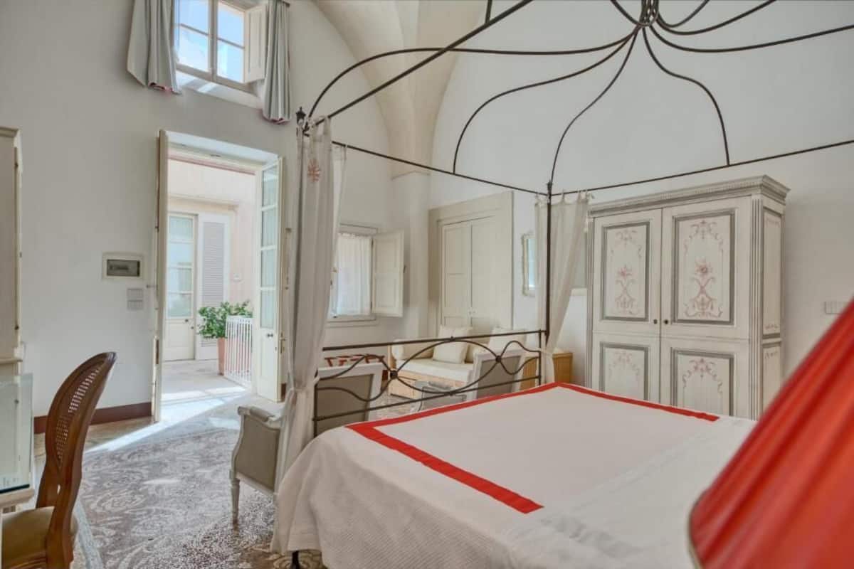A bedroom with a four-poster bed, a chair, a wardrobe, and a view of the outside.