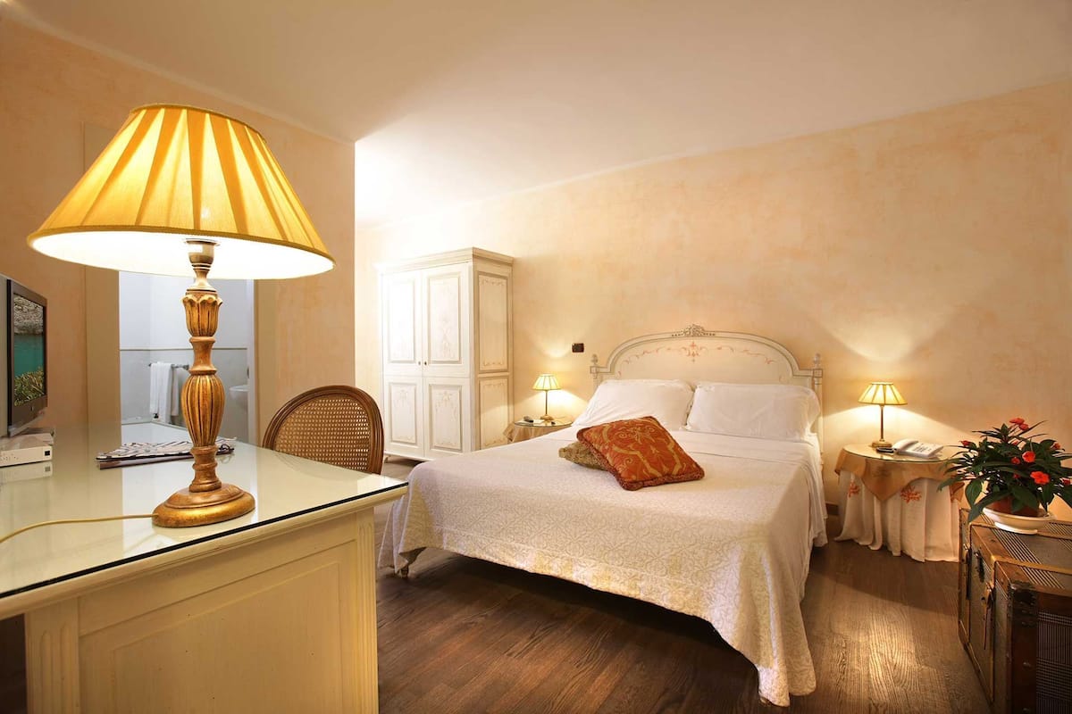 A hotel room with a bed, bedside lamps, a desk with a lamp, a chair, a wardrobe, and a small table with a plant.