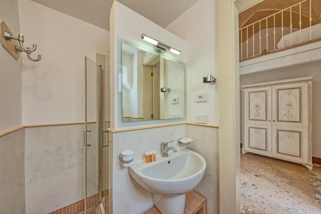 Un baño con cabina de ducha de vidrio, lavamanos con espejo y mueble blanco con detalles decorativos.