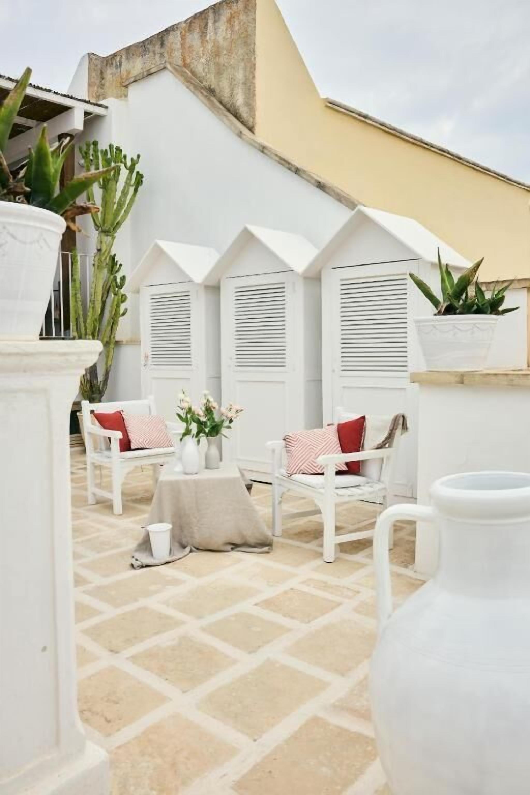 A white outdoor seating area with white chairs and tables, surrounded by potted plants and a white wall.