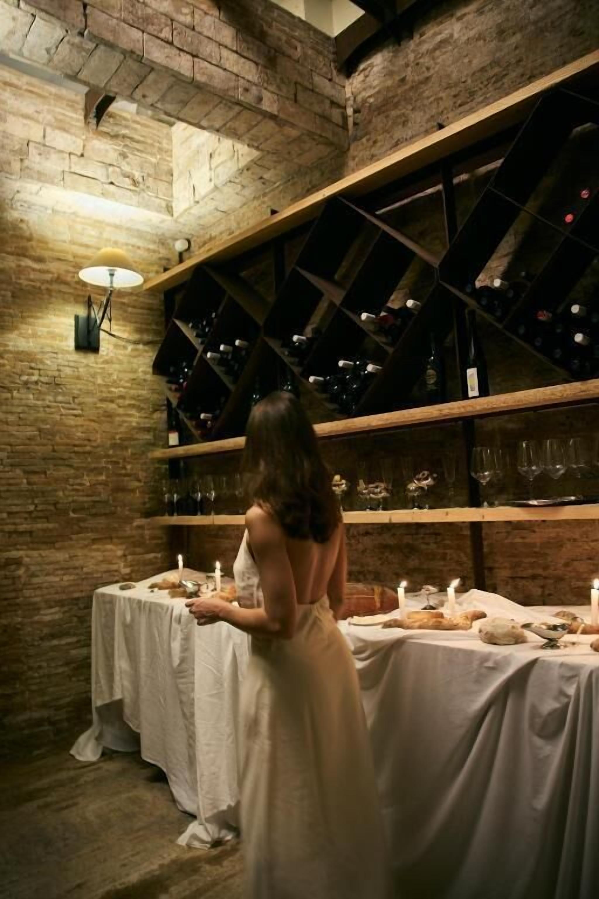 Una mujer con vestido blanco está de pie junto a una mesa con velas, en una habitación con paredes de ladrillo visto y vigas de madera.
