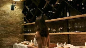 Una mujer con vestido blanco está de pie junto a una mesa con velas, en una habitación con paredes de ladrillo visto y vigas de madera.