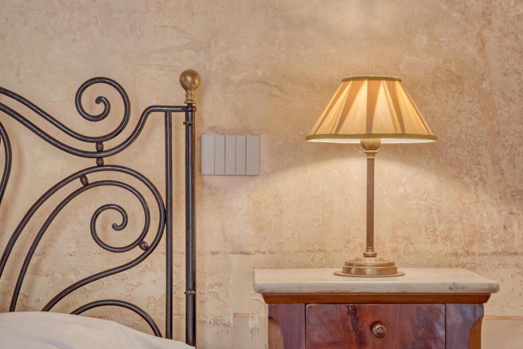 A bed with a metal headboard, a bedside table with a lamp, and a wall-mounted light switch.