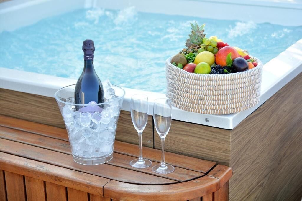 Een houten bank met een glas champagne, twee lege wijnglazen en een fruitmand bij een jacuzzi.