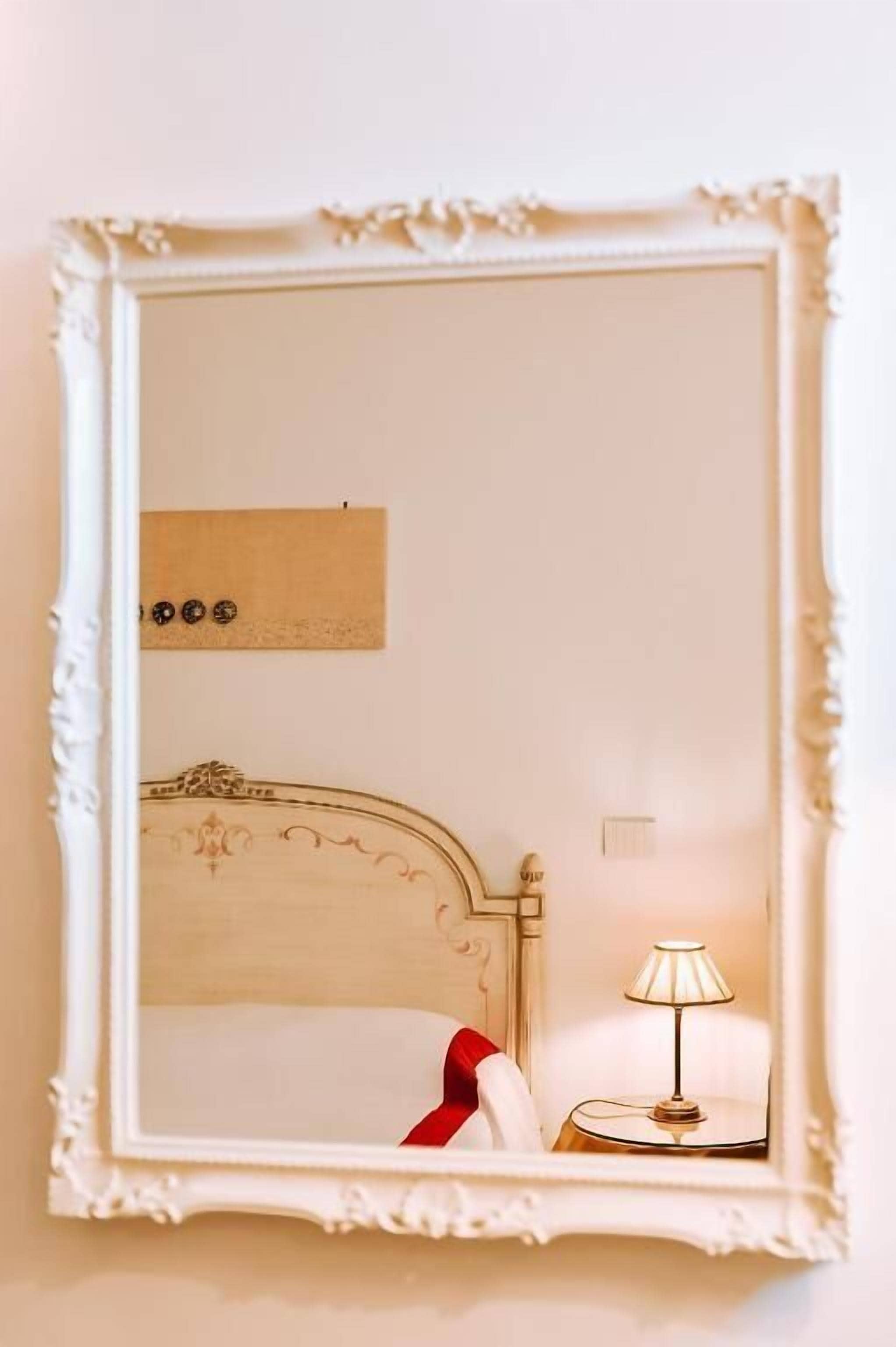 A mirror with a white ornate frame, a bed with a white headboard, a bedside lamp, and a wall-mounted light switch.