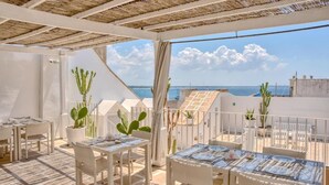 Eine Dachterrasse mit weißen Korbmöbeln, Topfpflanzen und Blick aufs Meer.