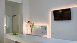 Een moderne badkamer met een wit wastafelmeubel, een bevestigde televisie en een spiegel die een douchegedeelte weerspiegelt.