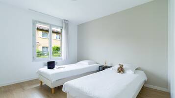A room with two single beds, a window with curtains, a small table with a lamp, and a teddy bear on one bed.