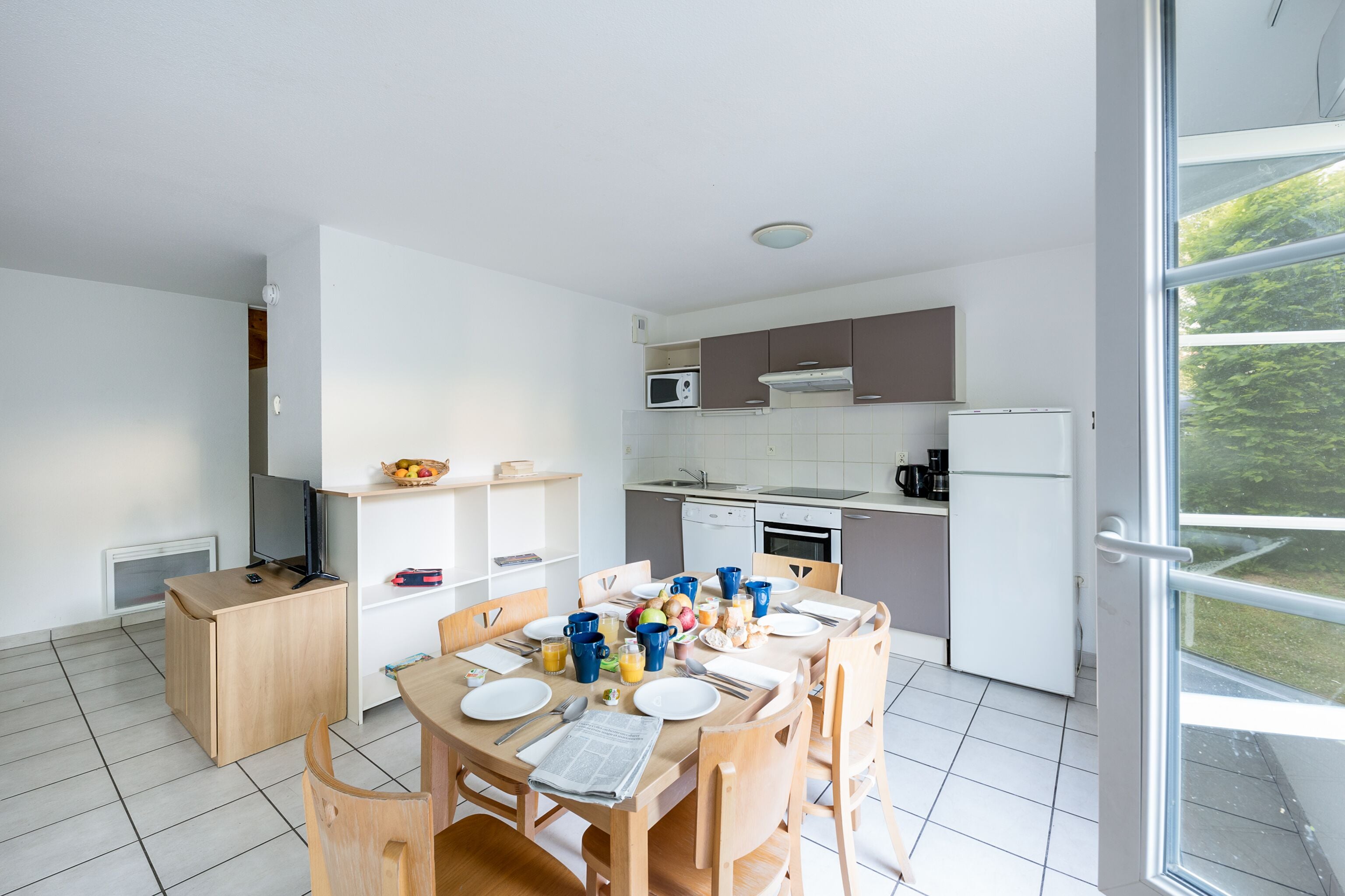 Een moderne keuken met witte kasten, een magnetron en een koelkast. Er staat een eettafel gedekt met borden, bekers en een krant.