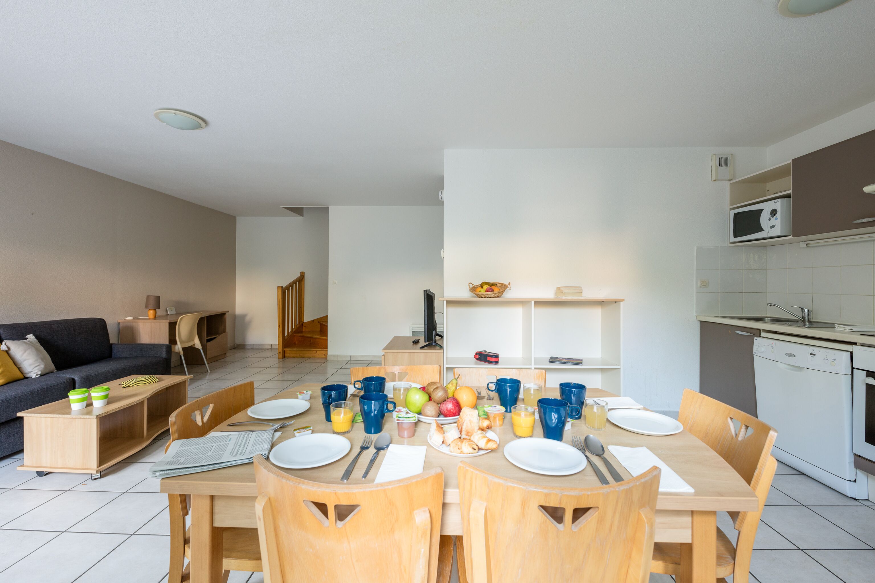 A modern living room with a sofa, coffee table, and TV. A dining area with a table set for four, chairs, and a kitchen with a microwave and sink.