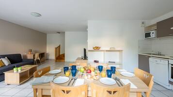 A modern living room with a sofa, coffee table, and TV. A dining area with a table set for four, chairs, and a kitchen with a microwave and sink.