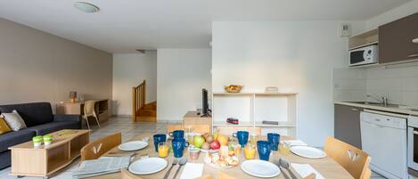 Uma sala de estar moderna com sofá, mesa de centro e televisão. Uma área de jantar com mesa posta para quatro, cadeiras e cozinha com micro-ondas e pia.