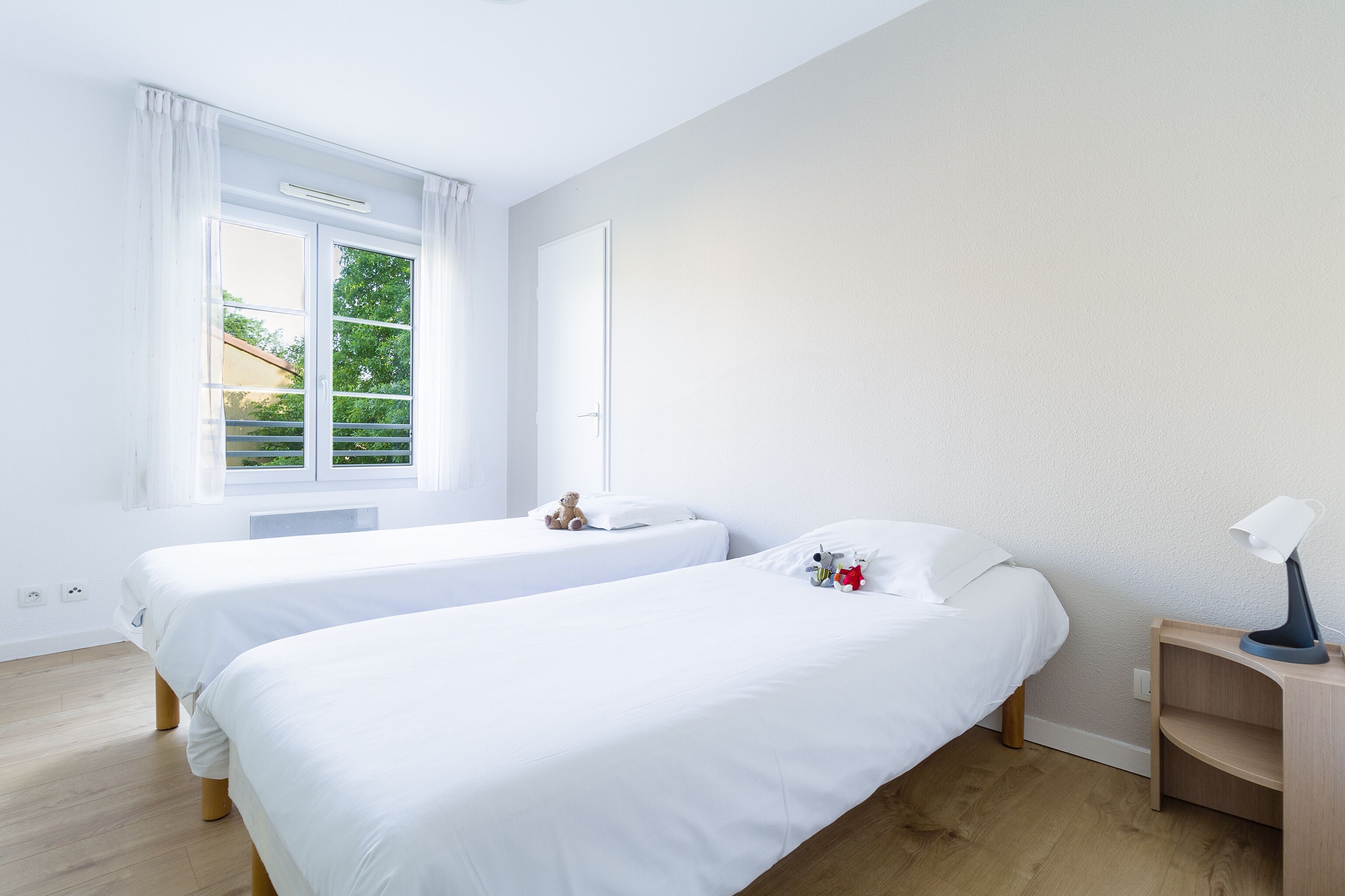 A room with two single beds, a window with curtains, a bedside table with a lamp, and a view of greenery outside.