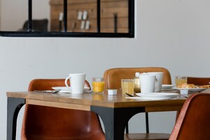 Mesa de desayuno dispuesta con platos, tazas y vasos, incluyendo jugo de naranja y té, en una habitación moderna con sillas de cuero y una mesa de madera.