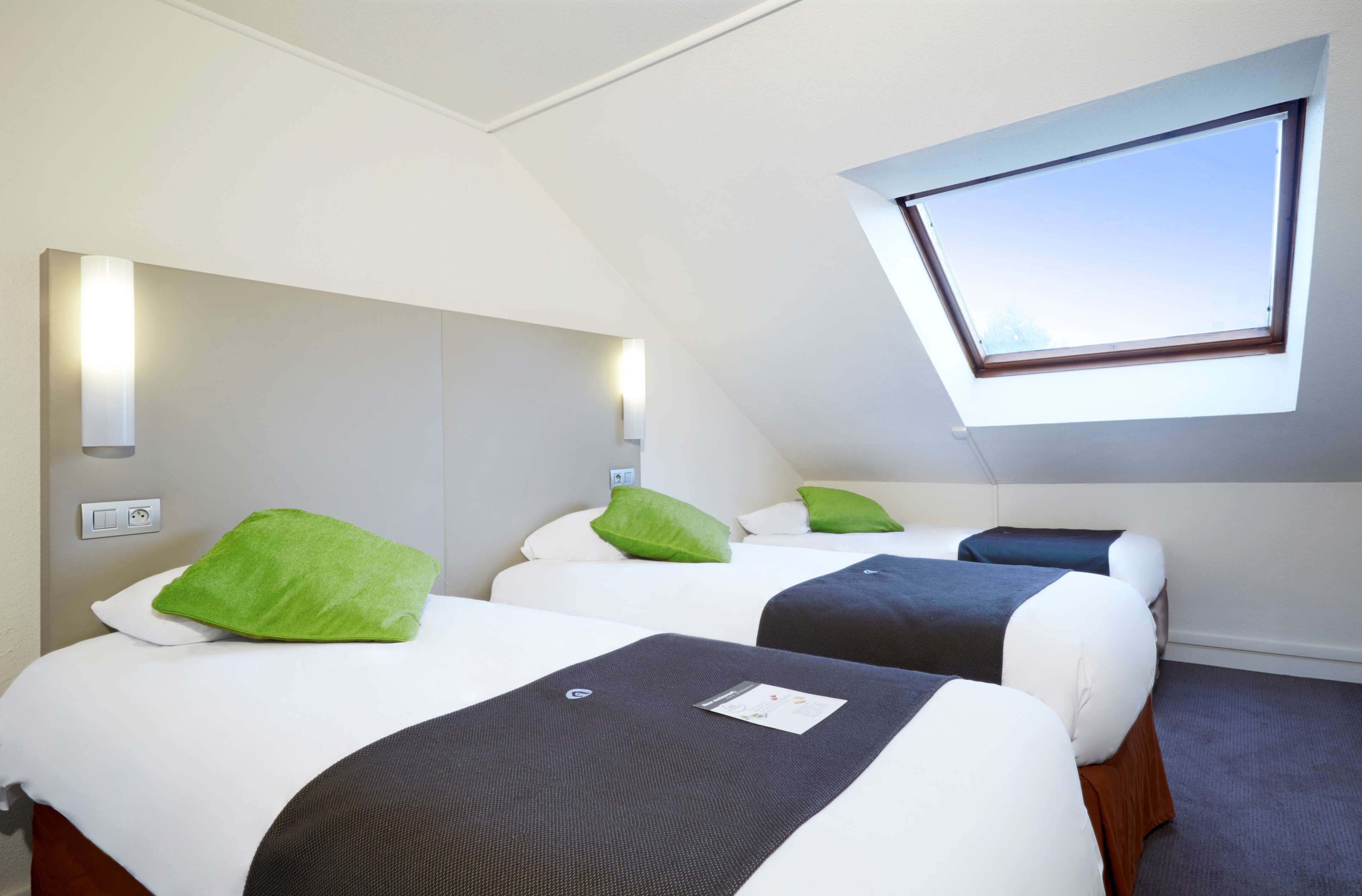 A hotel room with two beds, each with a pillow and a blanket. There is a skylight window above the beds, and a wall-mounted light fixture.