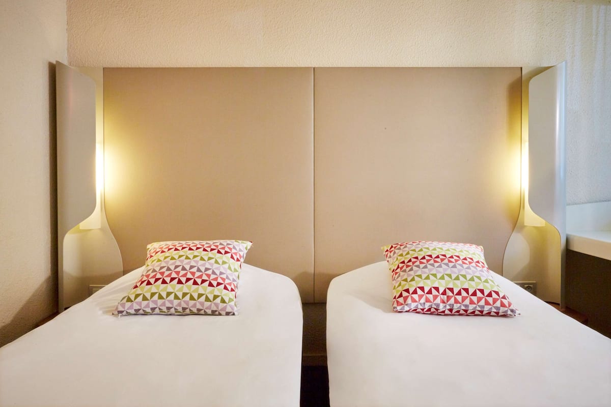 Two single beds with patterned pillows, a neutral-toned headboard, and wall-mounted lamps.