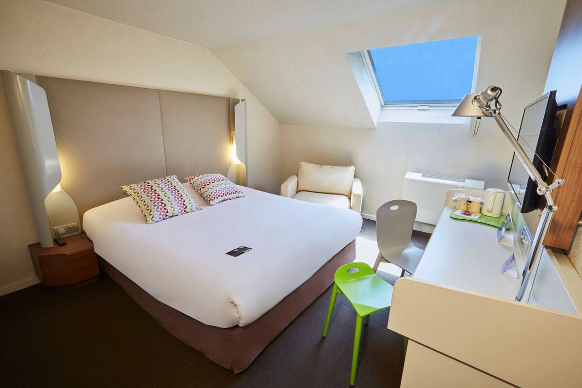 A hotel room with a bed, a desk, a chair, a nightstand, a lamp, a window, and a skylight.