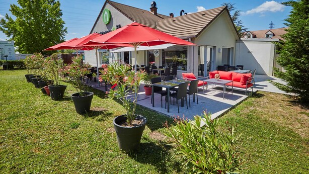 A patio area with outdoor seating, potted plants, and a building with a green logo. - Campanile Evry Ouest - Corbeil Essonnes (Corbeil-Essonnes)