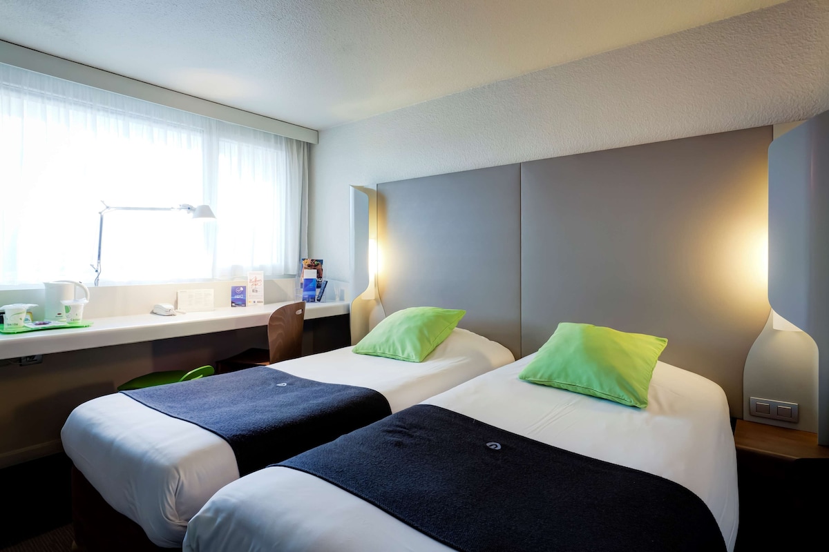 A hotel room with two beds, a desk with a lamp, a chair, and a window with curtains.