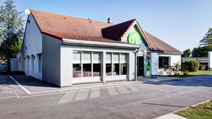 A modern building with a sloped roof, large windows, and a green logo on the facade. - Campanile Evry Ouest - Corbeil Essonnes (Corbeil-Essonnes)
