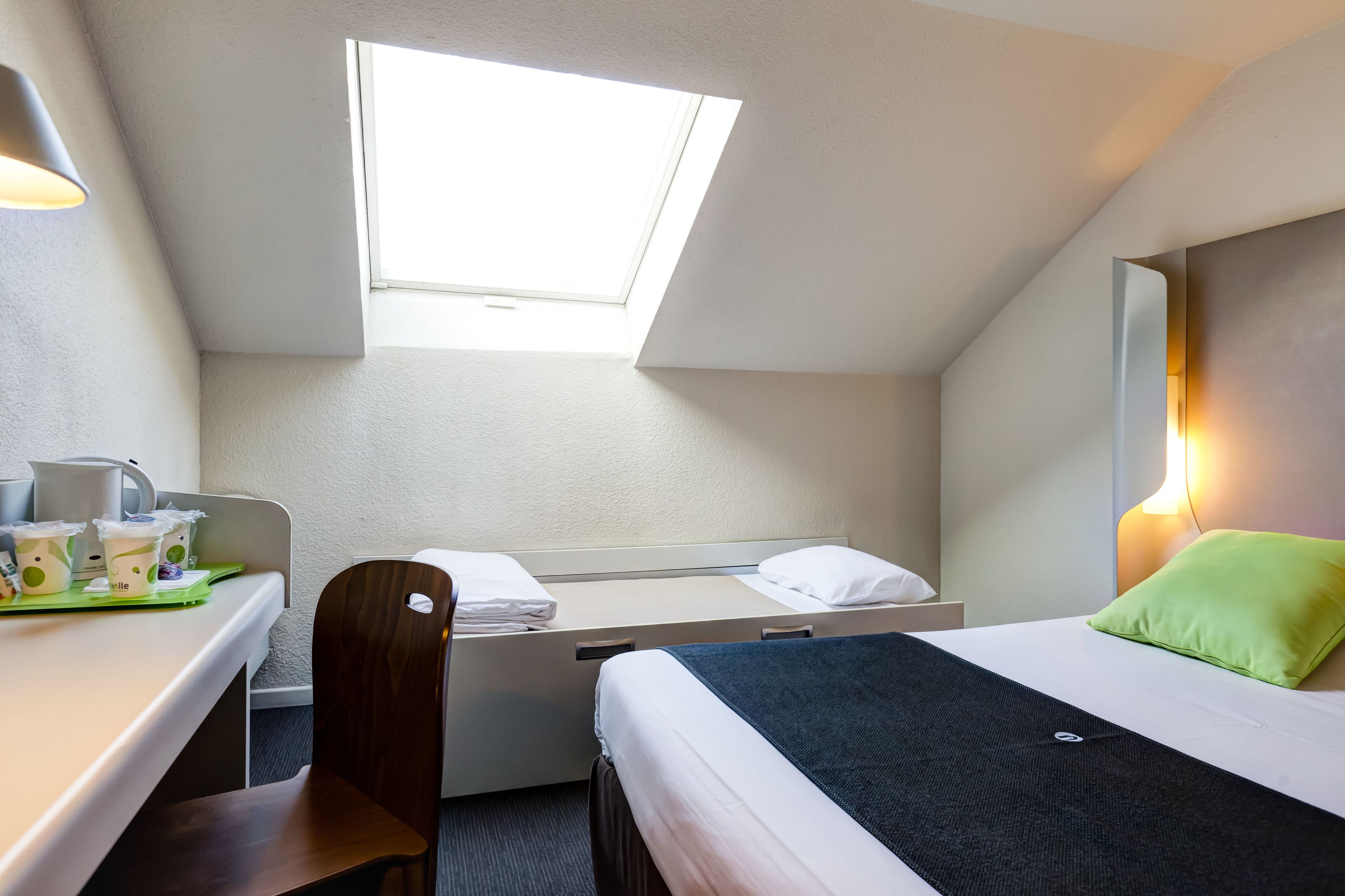 Une chambre d’hôtel avec une lucarne, un bureau avec une chaise, un lit avec un oreiller vert et une table de chevet avec une lampe.