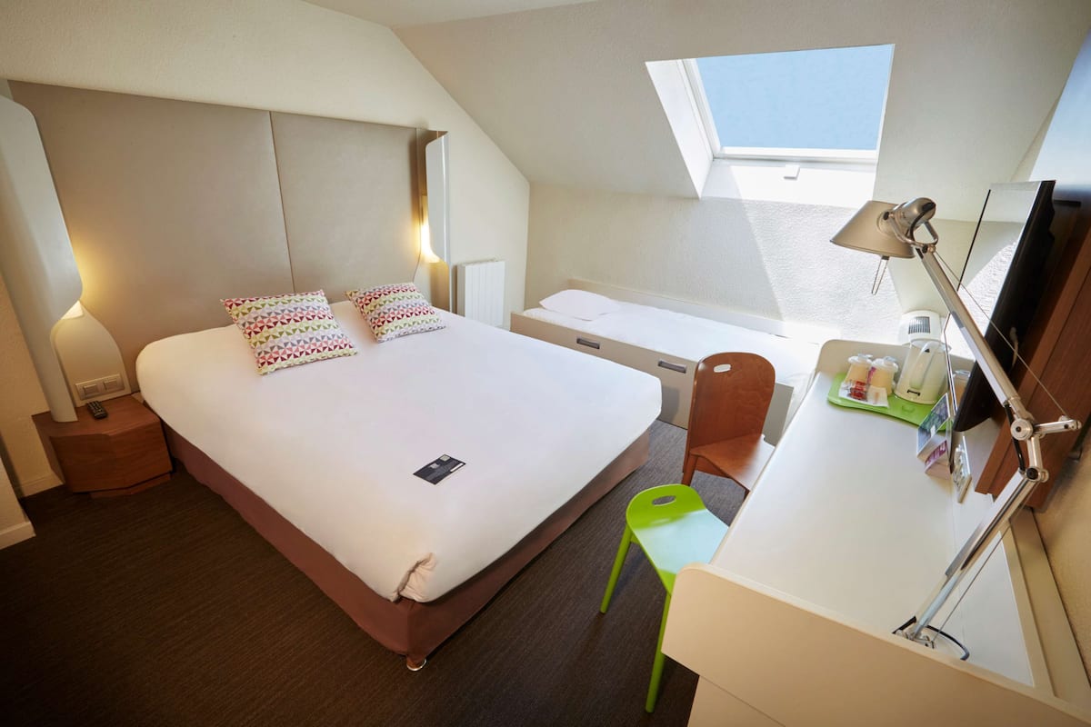 A hotel room with a bed, bedside table, desk, chair, and a skylight.