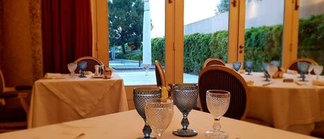 A dining area with tables set for a meal, featuring crystal glassware and a view of a garden through large windows.