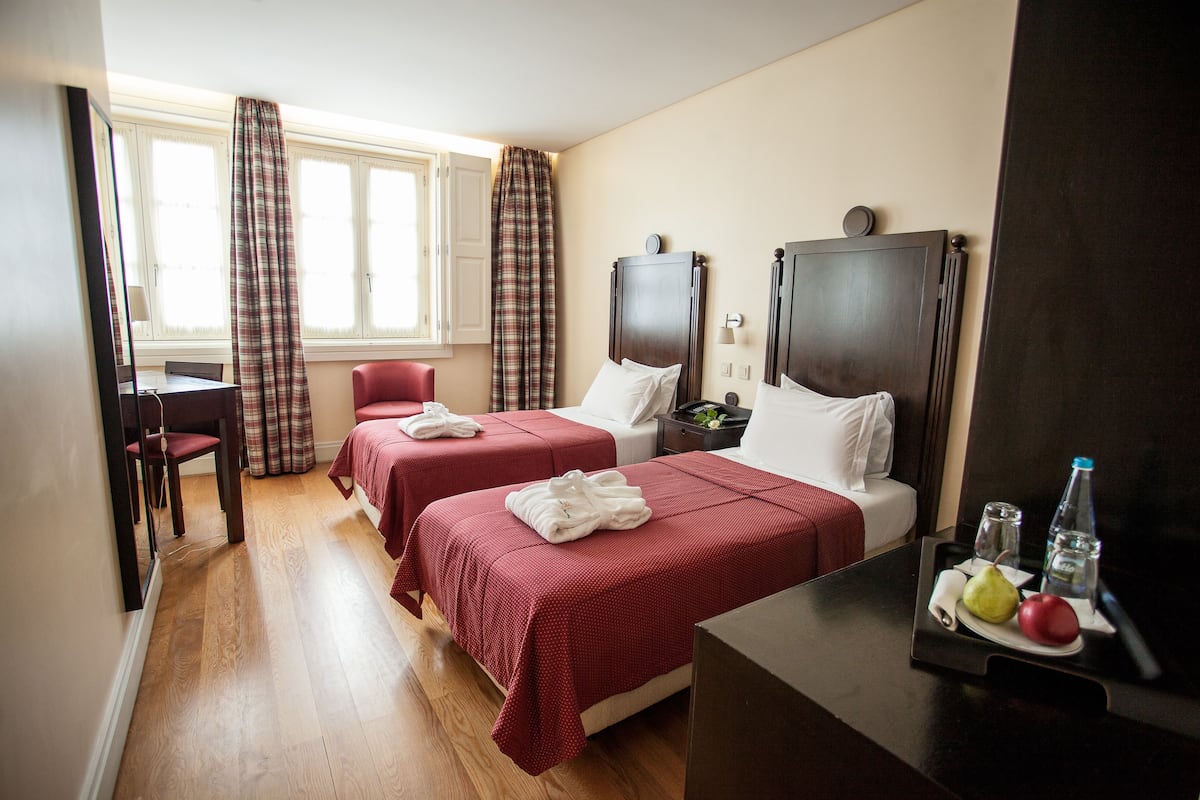 Habitación de hotel con dos camas, un escritorio y una silla. Tiene una ventana con cortinas, una mesita de noche con una botella y vasos, y un plato de fruta.
