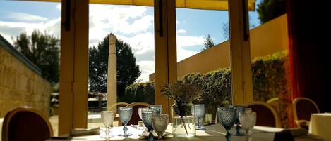 Un área de comedor al aire libre con una mesa preparada para dos, que incluye vasos de vidrio transparente y un jarrón con flores.