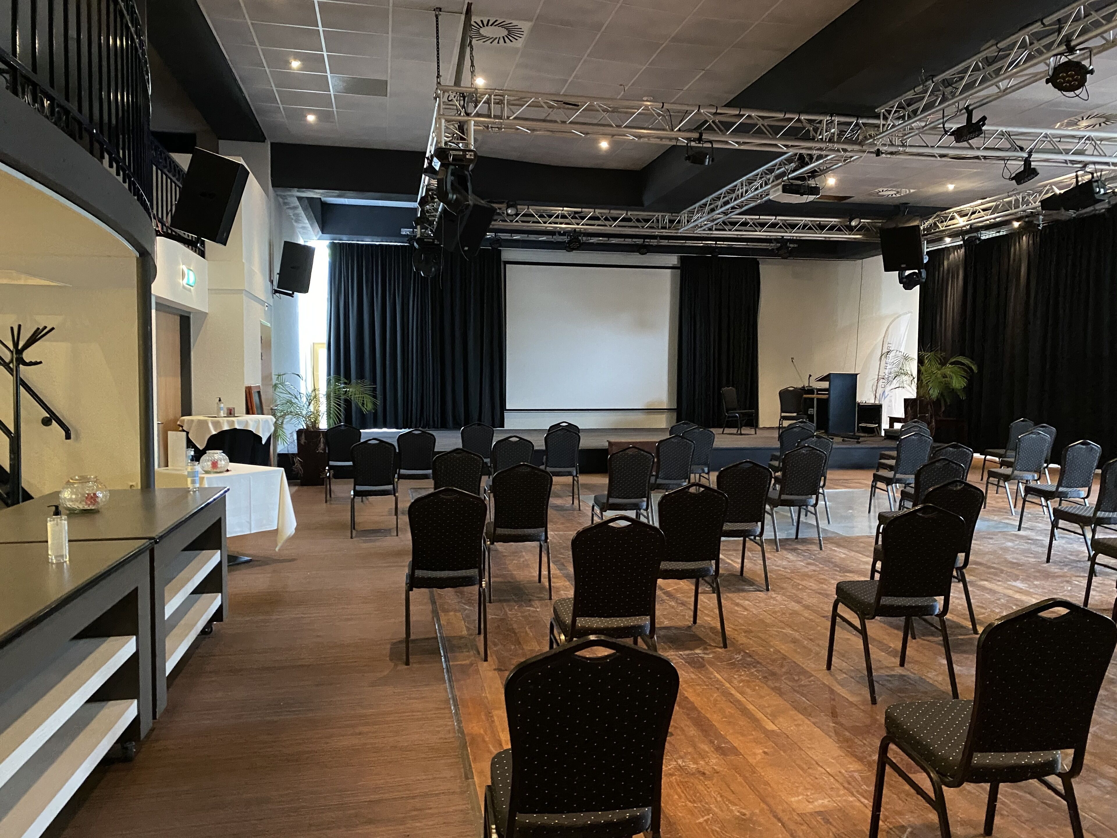 A conference room with rows of chairs, a stage with a projector screen, and a speaker system.