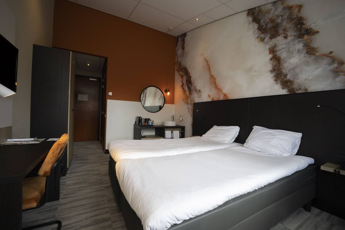 A hotel room with a large bed, a desk, a chair, a round mirror, a nightstand, and a wall with a marble-like design.
