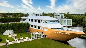 A modern houseboat with a large deck, glass-enclosed areas, and a garden with neatly trimmed bushes. - Fletcher Wellness-Hotel Brabant-Mill (Mill)