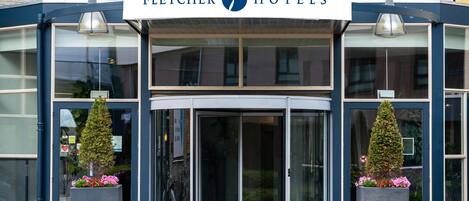 The Fletcher Hotels entrance with a glass facade, double doors, and two potted plants.