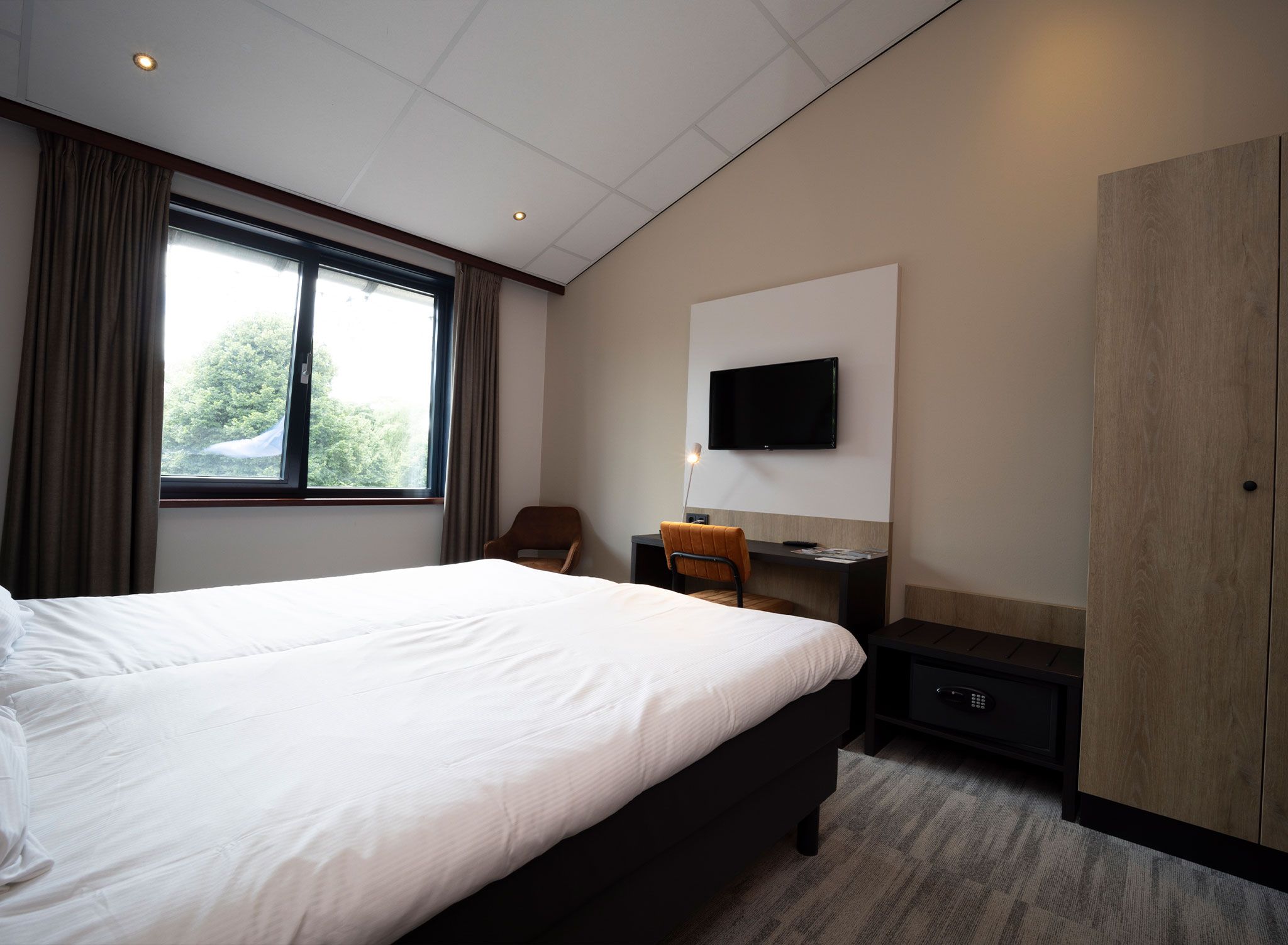 Ein Hotelzimmer mit einem großen Bett, einem Schreibtisch mit Stuhl, einem Wandfernseher und einem Kleiderschrank.