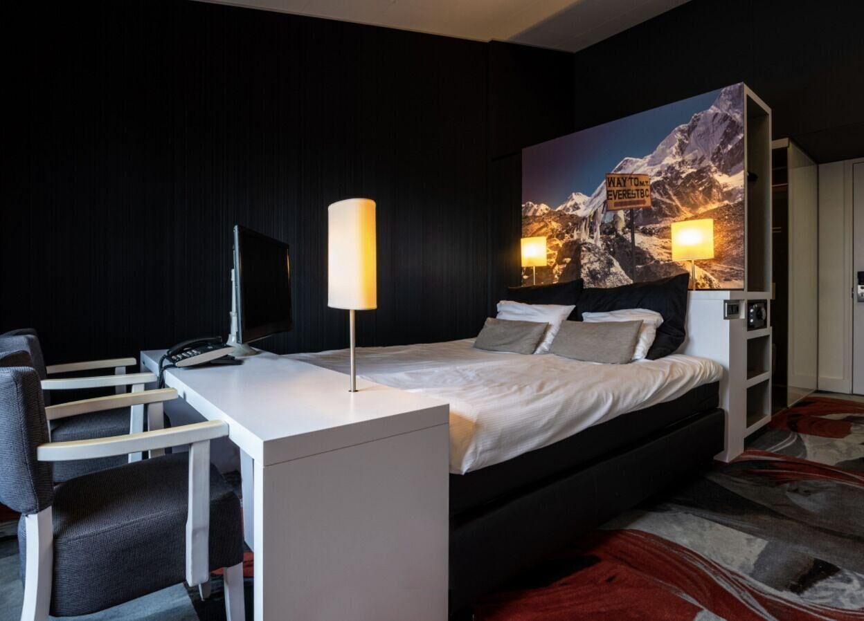 A modern hotel room with a bed, desk, chair, lamp, and a large mountain view painting.