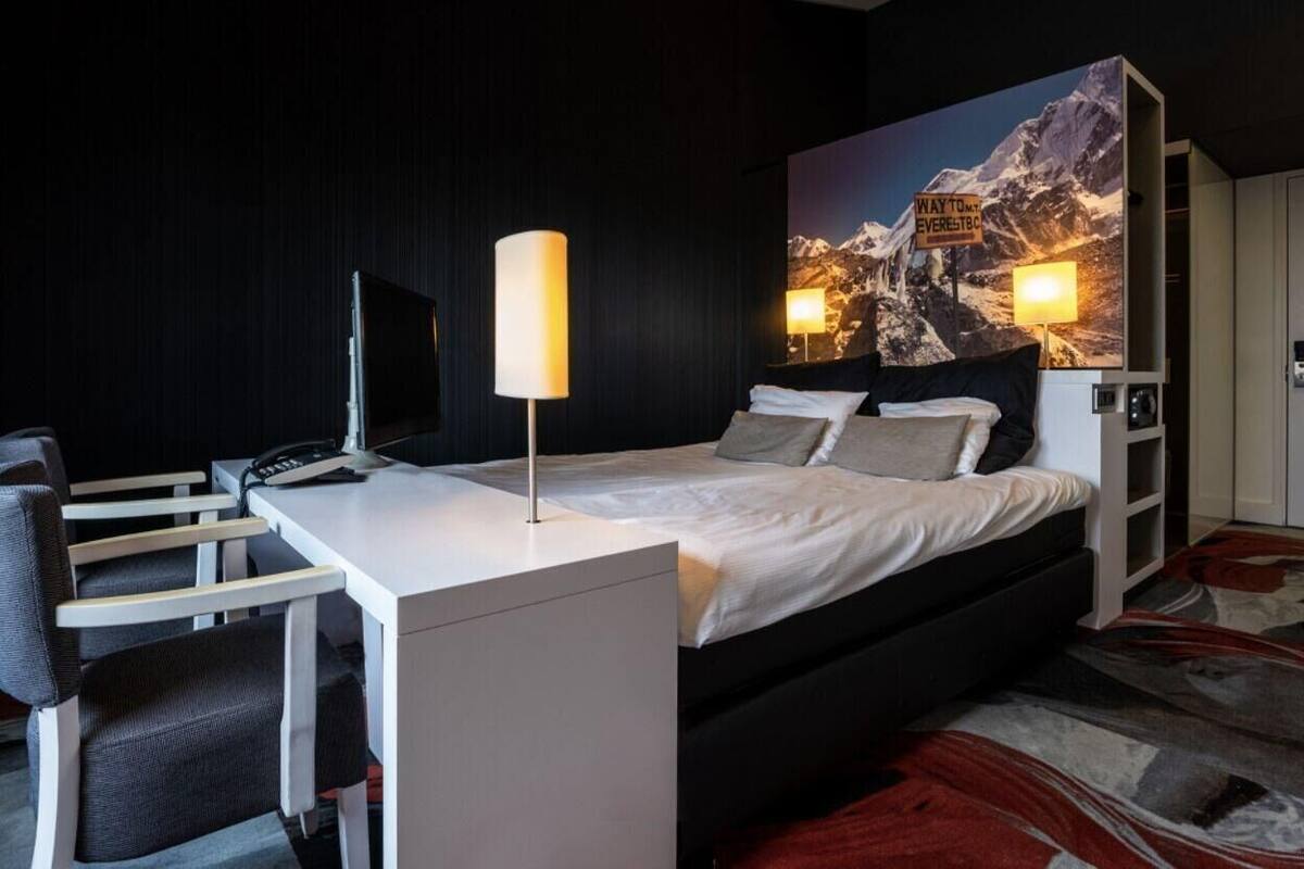 A modern hotel room with a bed, desk, chair, lamp, and a large mountain view painting.
