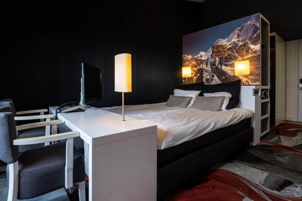 A modern hotel room with a bed, desk, chair, lamp, and a large mountain view painting.