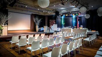 Un salón de conferencias con filas de sillas blancas, un escenario con un atril y una pantalla grande.