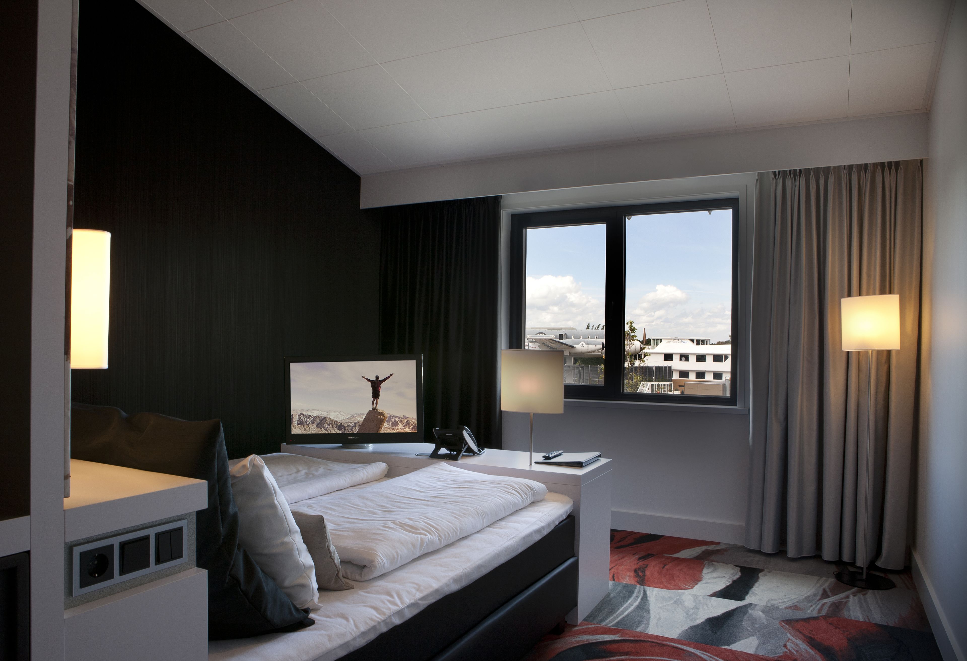 Habitación de hotel moderna con una cama grande, un escritorio, una silla, un televisor y una ventana con vistas a la ciudad.
