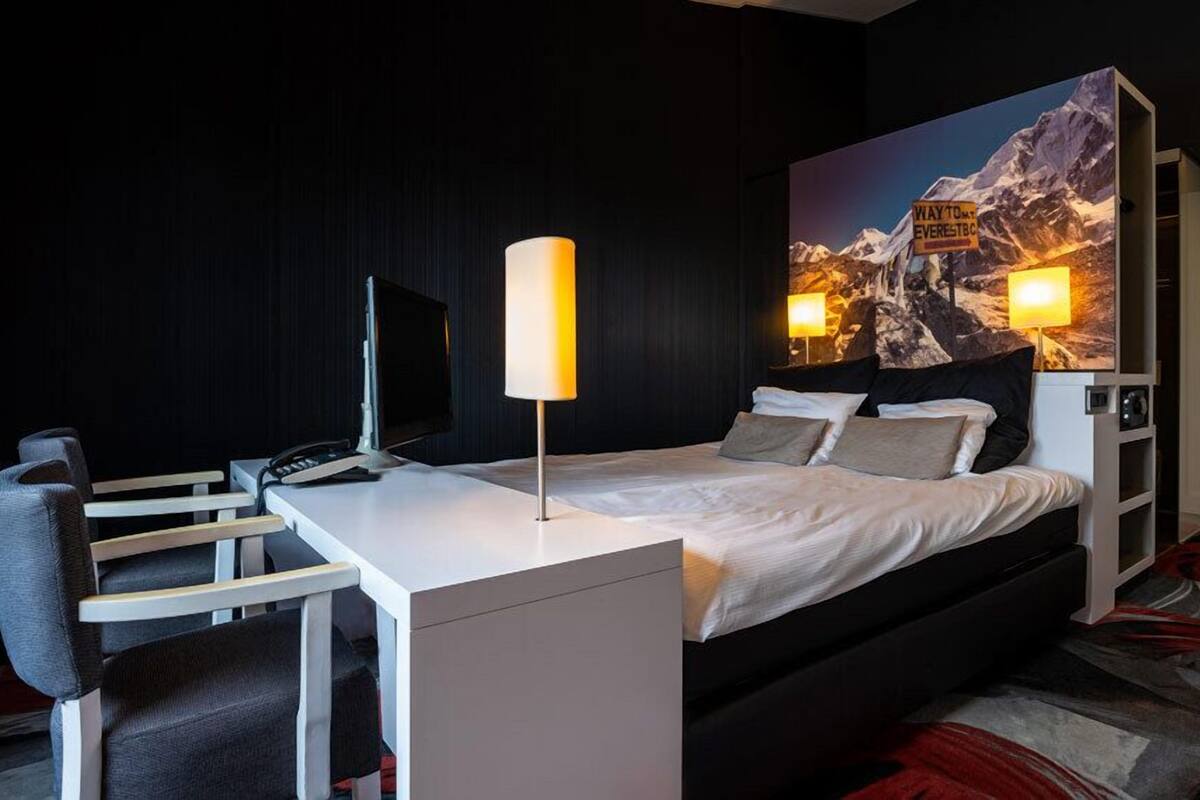 A modern hotel room with a large bed, a desk with a computer, and a chair. There is a painting of a mountain landscape on the wall.