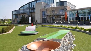 A modern building with a curved glass facade, a courtyard with artificial grass, a large copper bowl, and outdoor seating. - Fletcher Wellness-Hotel Brabant-Mill (Mill)