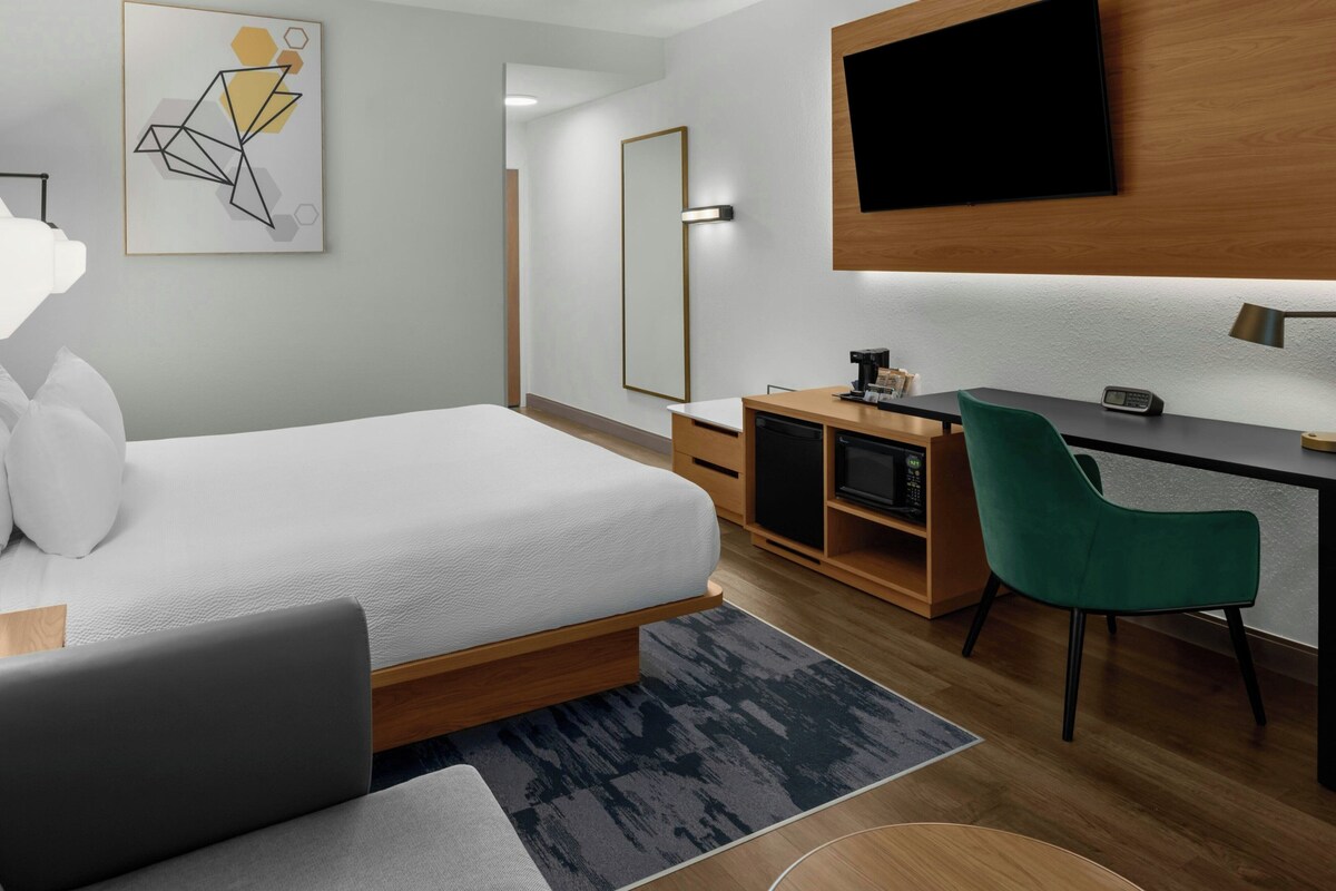 A modern hotel room with a bed, desk, green chair, flat-screen TV, and artwork on the wall.