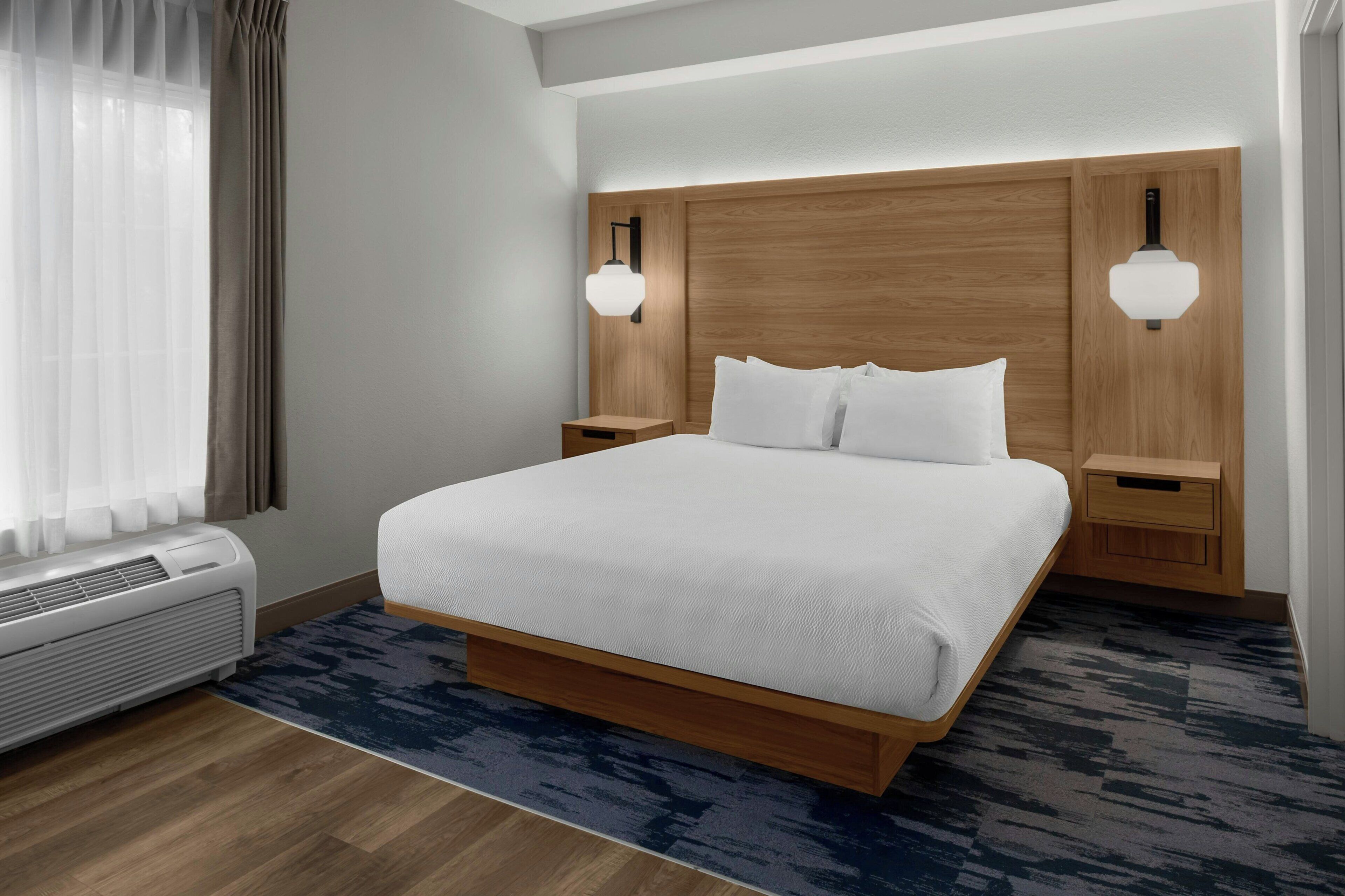 Una habitación de hotel con una cama grande, cabecero de madera, lámparas de noche y un ventanal con cortinas.