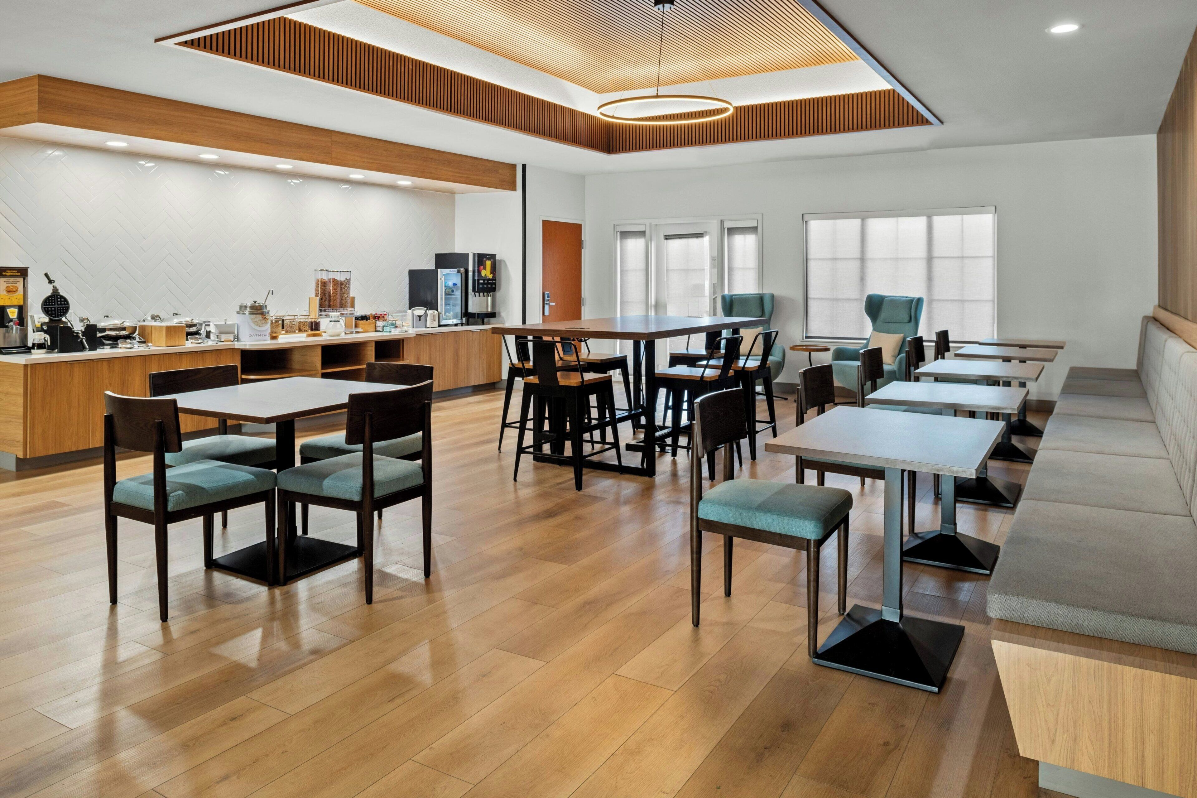 A modern hotel lobby with a wooden reception desk, multiple seating areas with tables and chairs, and a large window providing natural light.