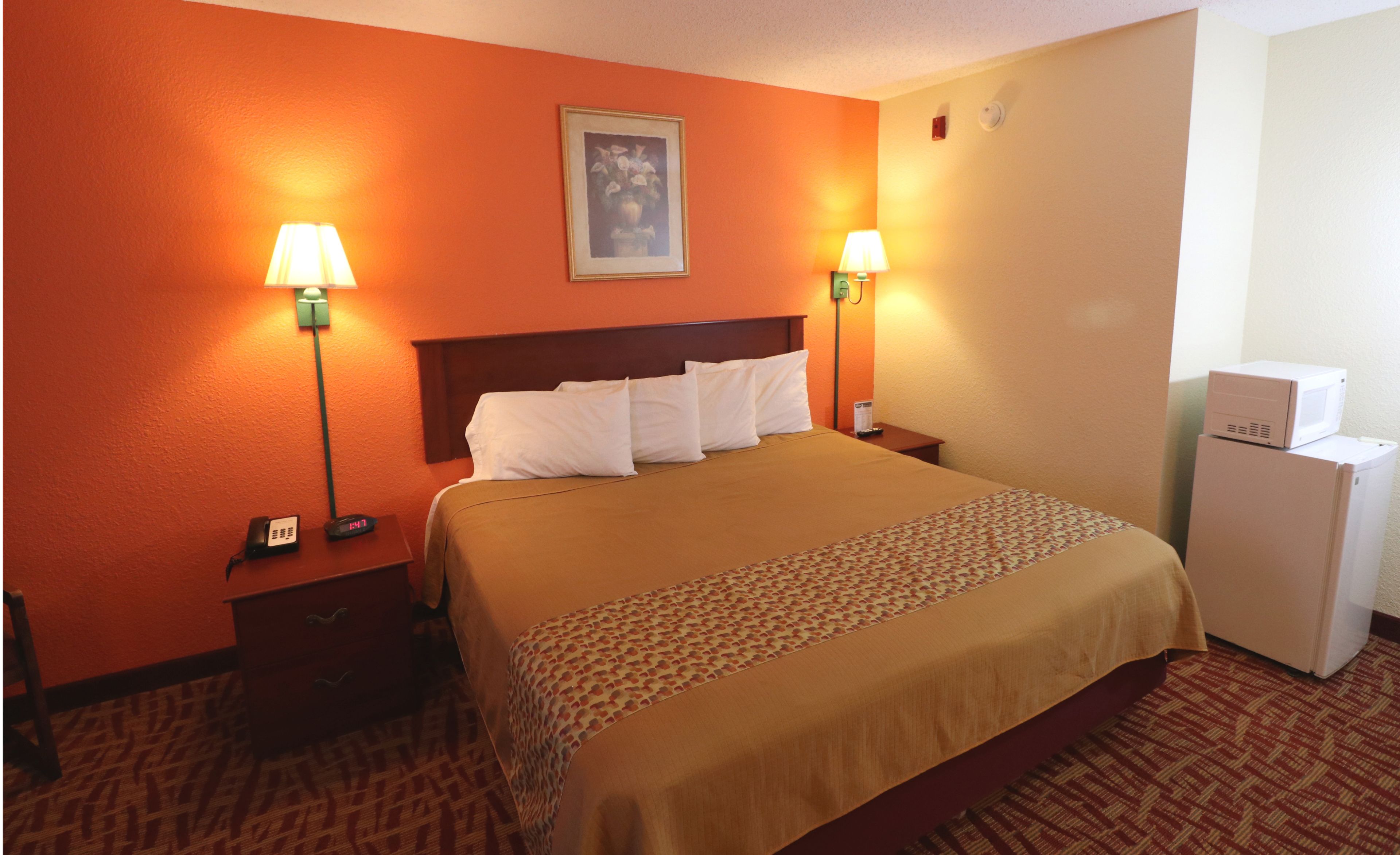 A hotel room with a bed, two bedside lamps, a nightstand, a clock, a framed picture on the wall, and a refrigerator.