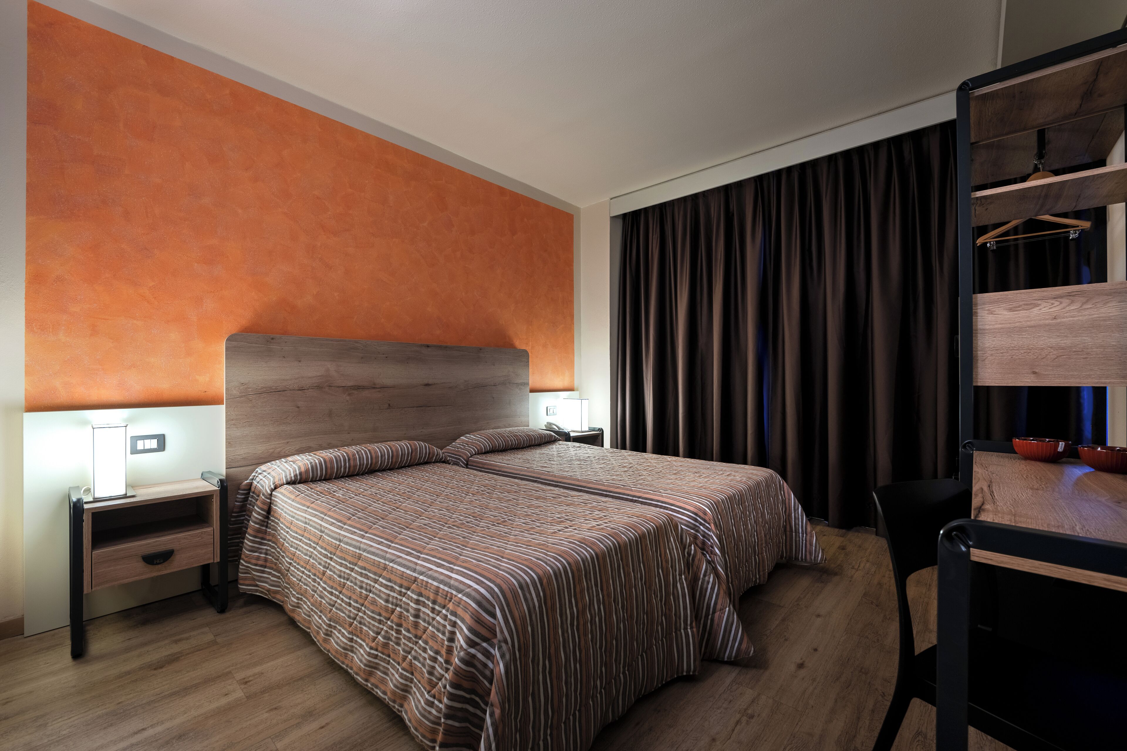 Habitación de hotel con cabecera de madera, cama con ropa de cama a rayas, mesita de noche con lámpara, escritorio con silla y armario con espejo.