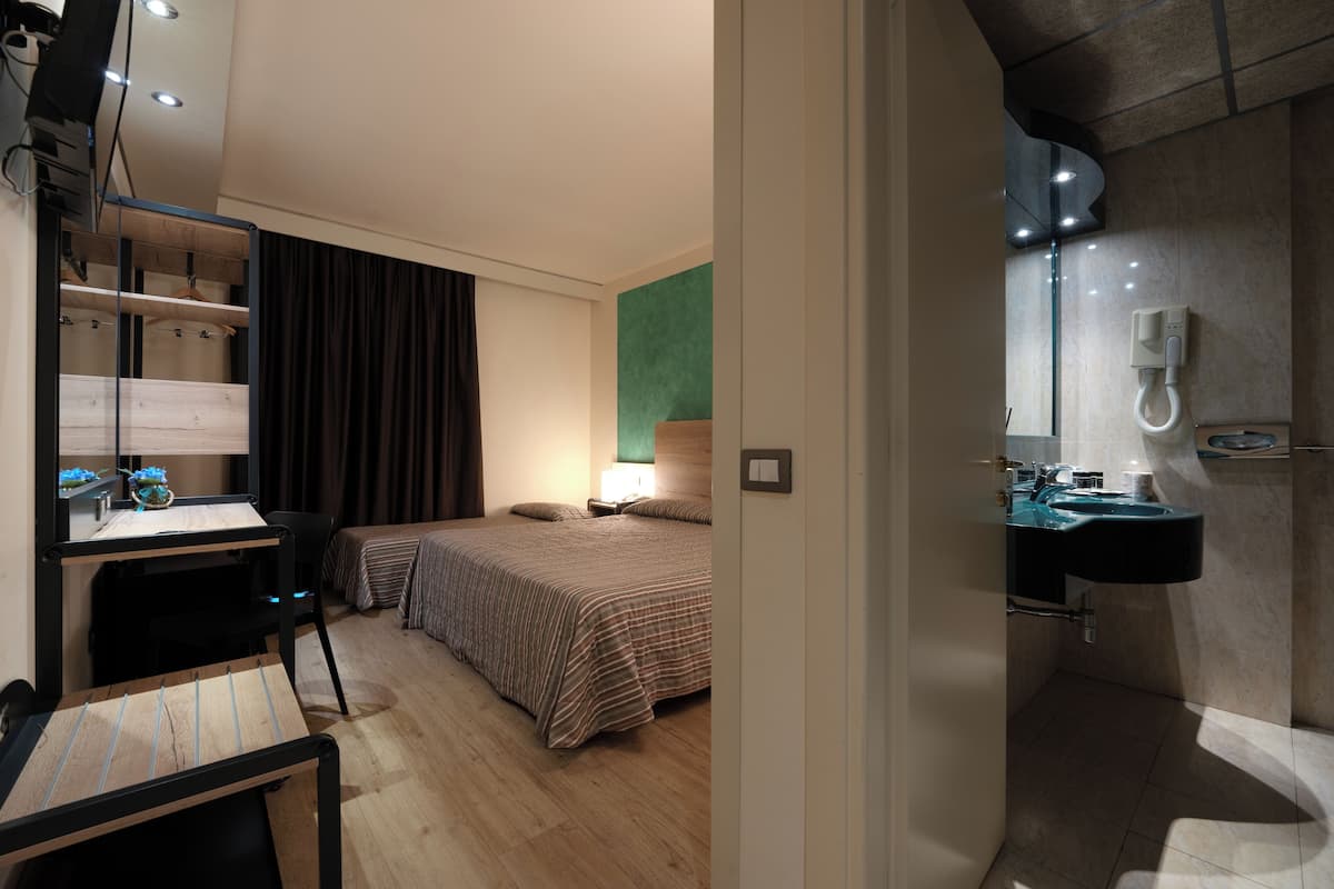 Habitación de hotel con cama, escritorio con silla, espejo, lavamanos y zona de ducha.