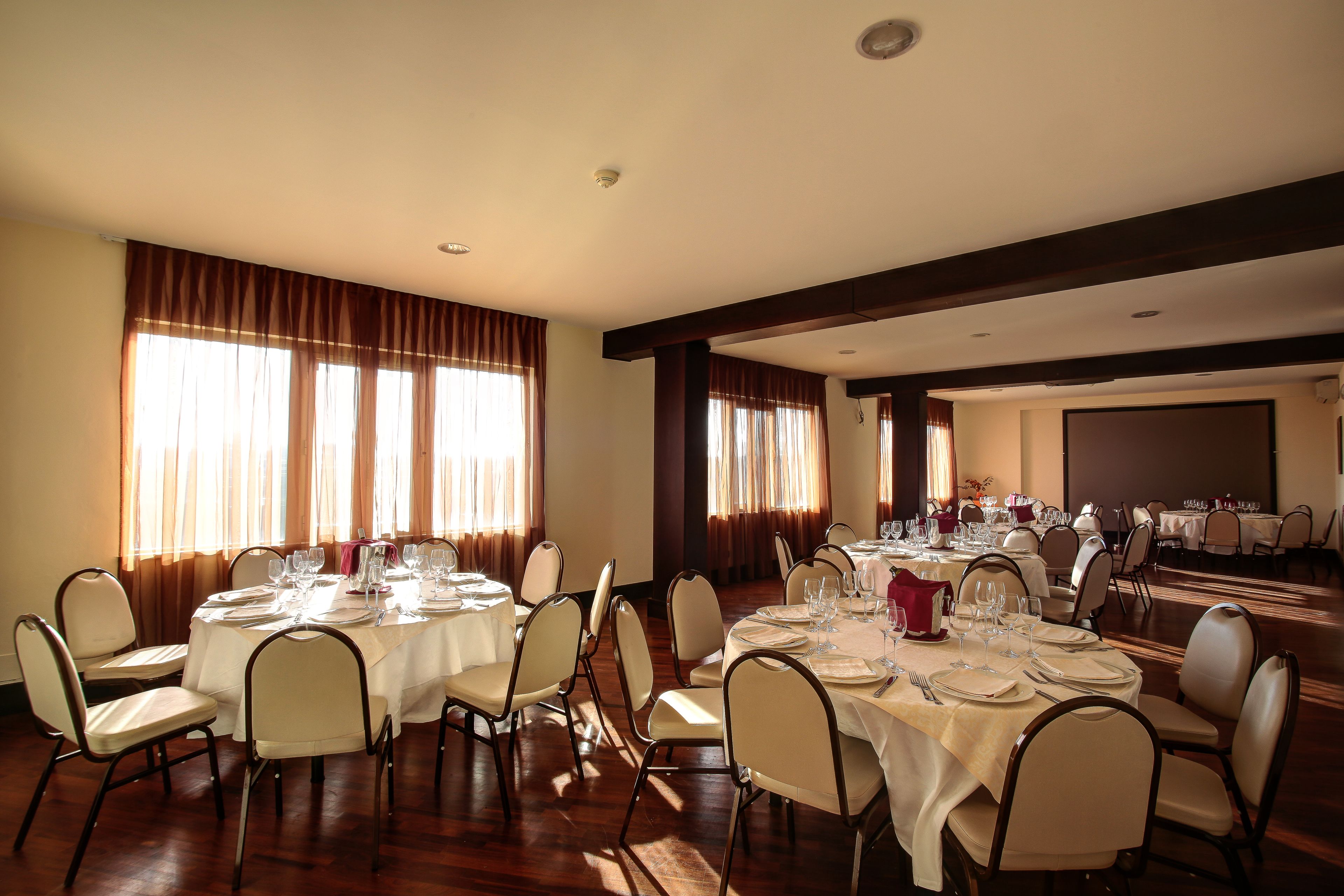 A banquet hall with round tables set for dining, large windows with curtains, and a stage area.