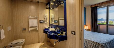 A modern bathroom with a sink, toilet, and bidet, a large mirror, and a bed with a view of the outdoors.